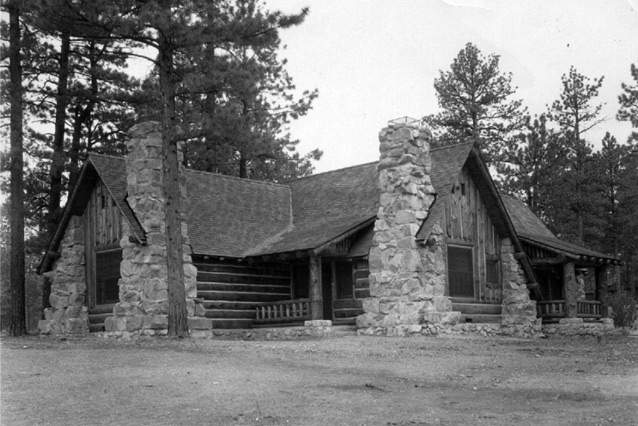 A low log cabin with a broad roof and prominent stone chimneys stands among pine trees in a black-and-white photograph.