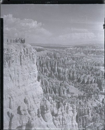 A small group stands at the upper left of a broad amphitheater overlook in a tall historic photograph.