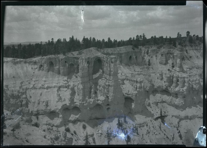 A historic scan shows a wooded rim over a dense rock wall with dark openings; scratches and a pale blemish remain visible.