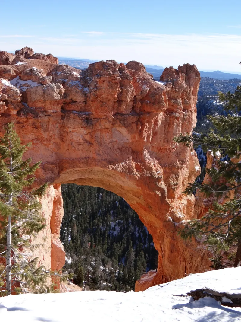 Vertical photograph of Natural Bridge: a red rock arch with a dark, forested landscape visible through its opening and patches of snow on surrounding rock.